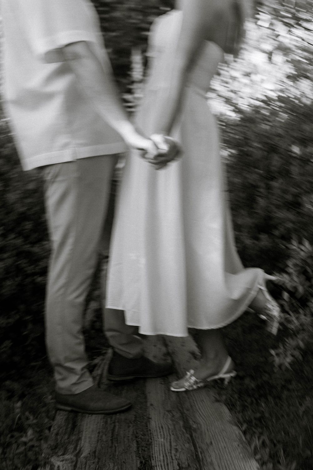 Black and white blurred photo of a couple holding hands in white dress and casual attire on a wooden deck.
