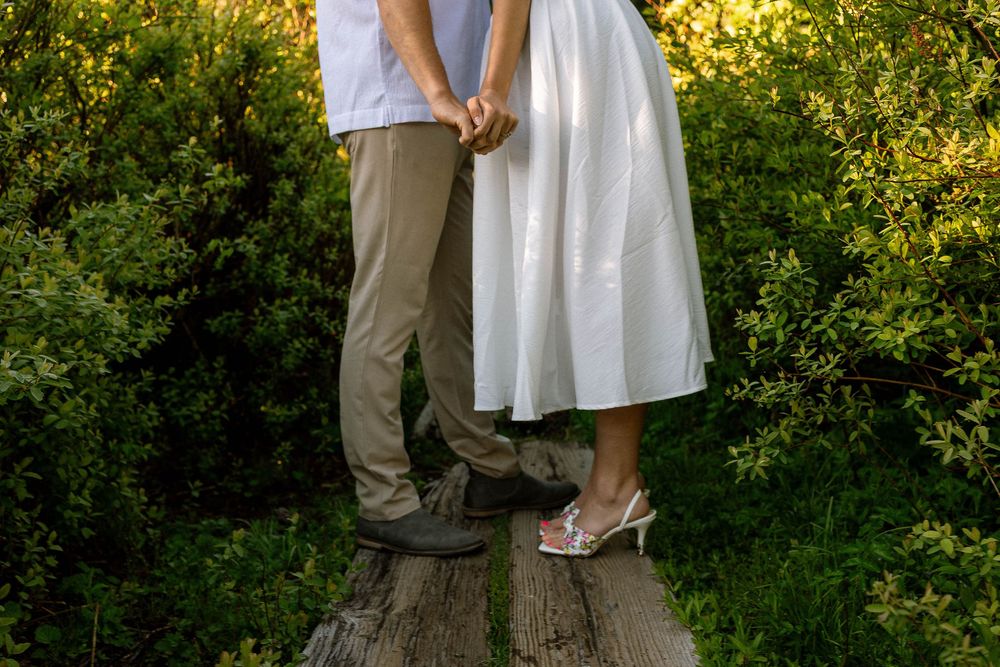 Couple holding hands in a garden, woman in white dress and heels, man in khaki pants, standing on wooden boards.