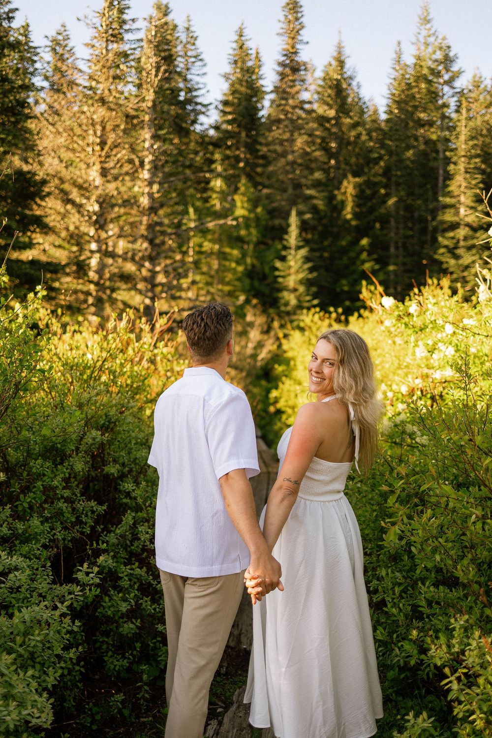 Couple holding hands in a sunlit forest meadow, woman in white dress smiling back at camera.
