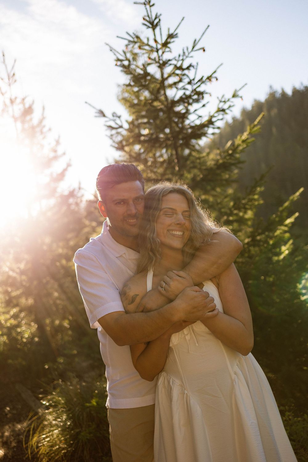 Couple embracing in golden sunlight among evergreen trees, man hugging woman in white dress from behind.