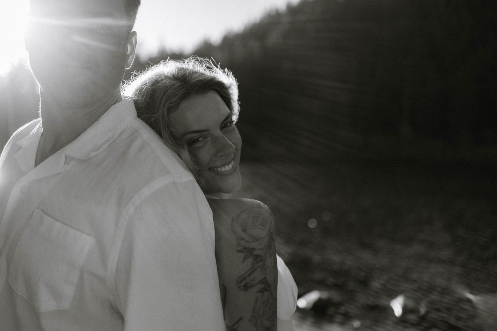 Black and white photo of a smiling woman with tattoos embracing a man in sunlight.