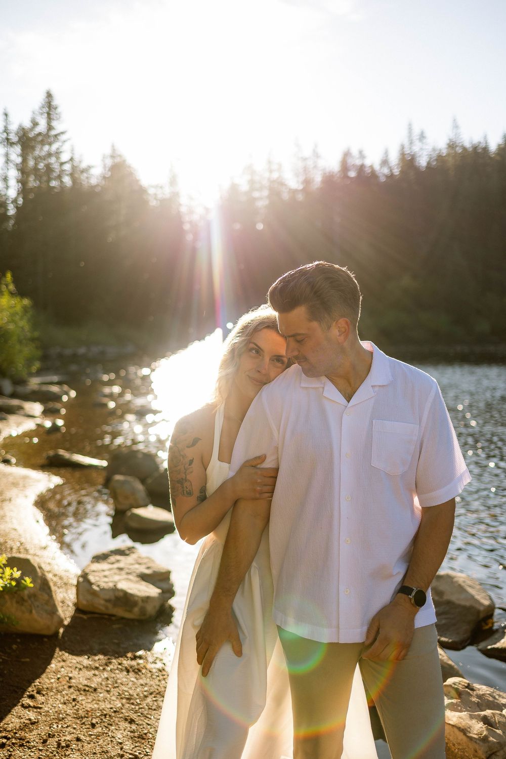 Couple embracing by a sunlit mountain lake surrounded by pine trees, golden hour light creating a romantic lens flare effect.