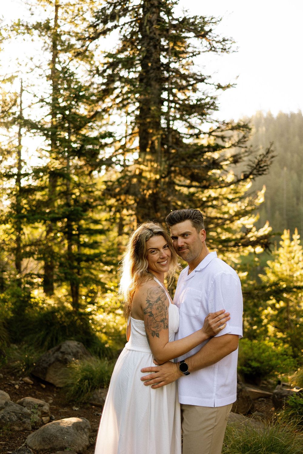 Couple embracing in golden forest light, woman in white dress with tattoo, man in white shirt and khakis.