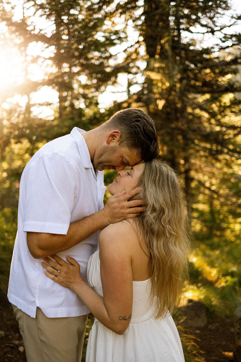 Couple sharing a romantic almost-kiss at golden hour in a forest, man in white shirt, woman in white dress.