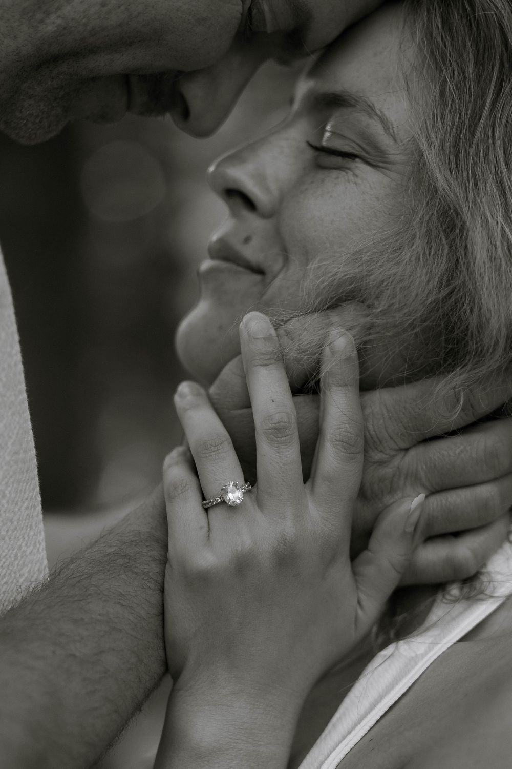 Close-up black and white photo of couple almost kissing, woman's hand with engagement ring touching man's face.