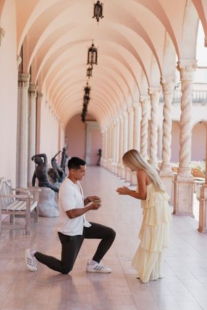 Man proposing on one knee to woman in yellow dress under arched colonnade.