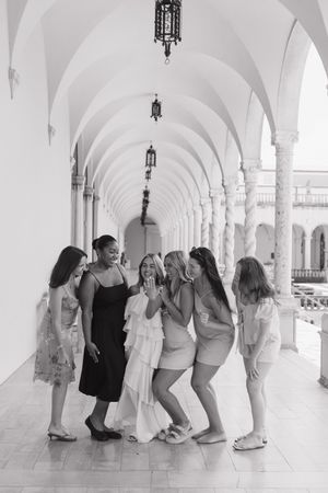 Black and white photo of six women laughing together in an elegant arched corridor.
