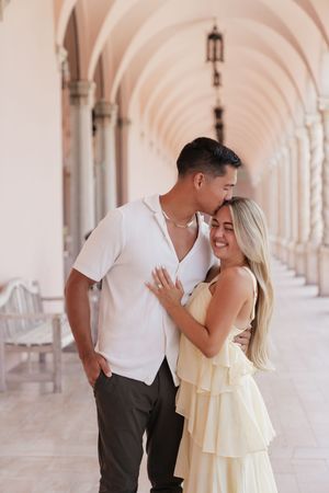 Couple sharing a tender kiss in a beautiful arched hallway, woman showing engagement ring.