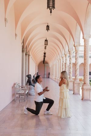 Man proposes on one knee to woman in yellow dress under pink arched colonnade.