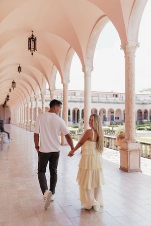 Couple walking hand in hand through a stunning pink arched colonnade overlooking a courtyard garden.