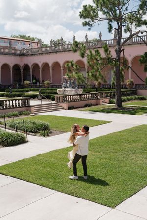Couple embracing in a romantic garden courtyard of a pink Mediterranean-style building with arched colonnades.