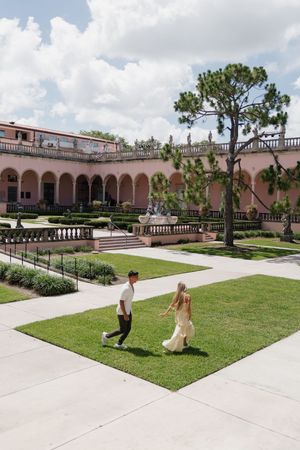 Couple walking hand in hand across lush green lawn at elegant pink Mediterranean-style courtyard with arched colonnades.