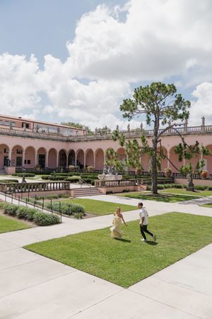 Couple running hand-in-hand across green lawn at elegant pink Mediterranean-style courtyard on sunny day.