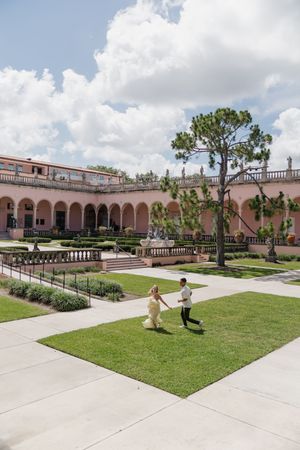 Joyful couple running hand in hand across lush green lawn of a grand pink Mediterranean-style estate.