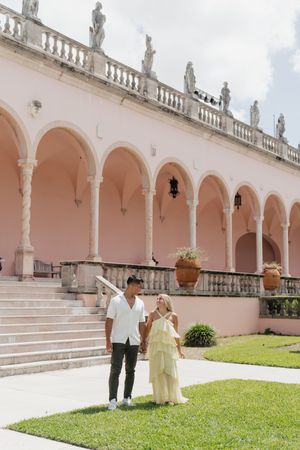 Couple holding hands and walking in courtyard of pink Mediterranean-style building.