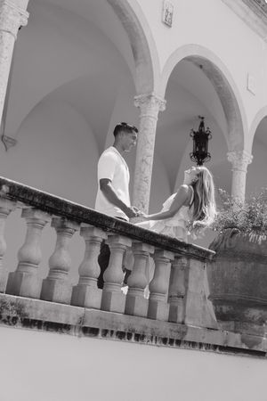 Black and white photo of couple holding hands on ornate balcony with arched architecture.