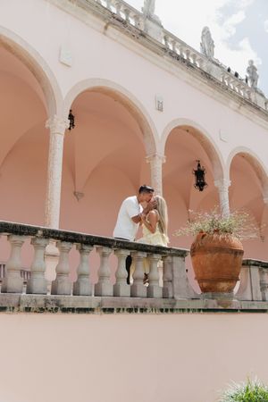 Couple sharing a kiss on a balcony with pink arched architecture and a large terracotta planter.