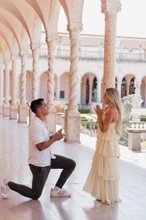 Man on one knee proposing to woman in yellow dress in elegant arched courtyard.