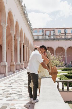 Engaged couple sharing kiss leaning against balustrade in pink courtyard arcade.