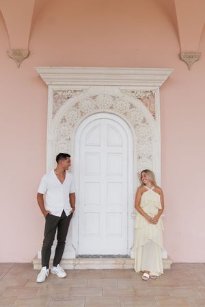 Couple standing apart by ornate white arched door on pink building exterior.