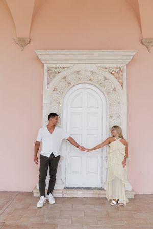 Newly engaged couple holding hands before ornate white door on pink wall.