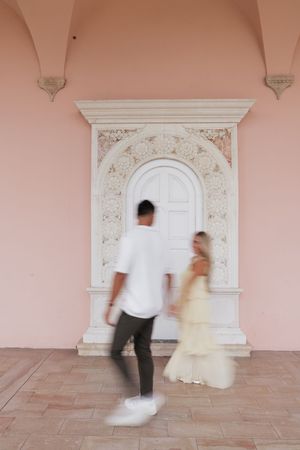 Blurred motion shot of couple walking toward ornate white arched doorway on pink wall.