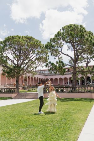 Man spinning woman in yellow dress on green lawn outside pink Mediterranean estate.