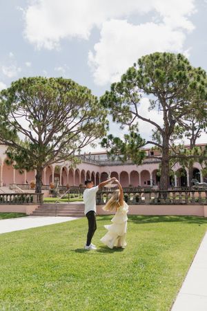 Couple dancing joyfully on green lawn in front of pink arched Mediterranean building.