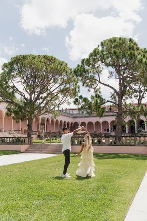 Couple dancing and twirling on green lawn in front of a pink arched building with tall trees.