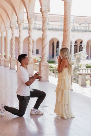 Romantic marriage proposal with man on one knee holding ring box for blonde woman in yellow dress.