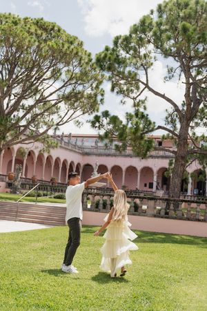 Happy couple dancing on green lawn with pink Mediterranean-style building in background.