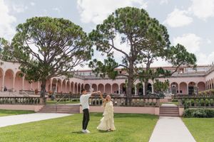 Couple dancing on green lawn at Ringling Museum courtyard with pink arched building and tall pine trees backdrop.