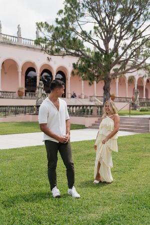 Couple sharing a relaxed conversation on green lawn outside pink Mediterranean estate.