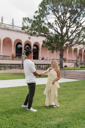 Couple holding hands on green lawn outside pink arched building, woman in yellow ruffled dress, man in white shirt.