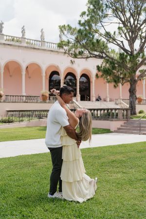 Couple embracing and kissing on green lawn outside pink Mediterranean-style building.