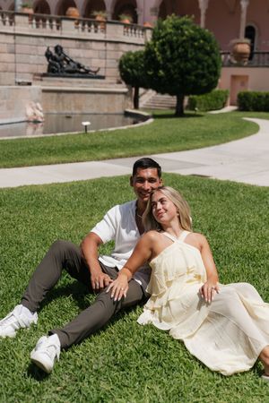 Couple sitting together on lush green grass in front of an elegant fountain and sculpted garden landscape.
