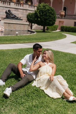 Couple sitting on green grass gazing at each other near a pink Mediterranean-style building.
