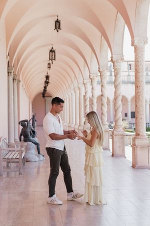 Couple holding hands in elegant pink archway colonnade with marble columns and hanging lanterns.