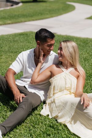 Smiling engaged couple sitting together on green grass, woman touching mans face tenderly.