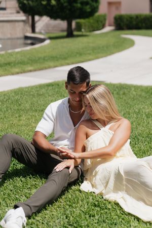 Couple sitting on lush green grass holding hands near a pink Mediterranean-style estate building.