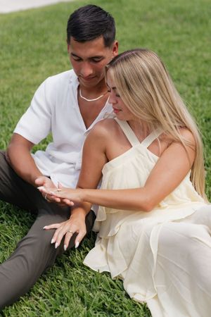 Newly engaged couple sitting on grass admiring engagement ring on womans hand together.