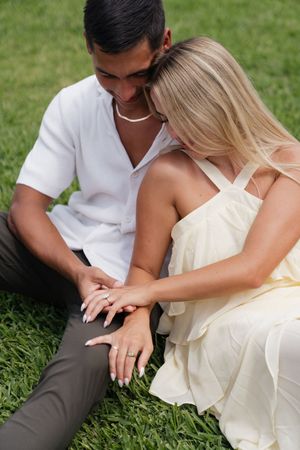 Couple sitting on green grass, woman in yellow dress showing engagement ring, man in white shirt leaning close.