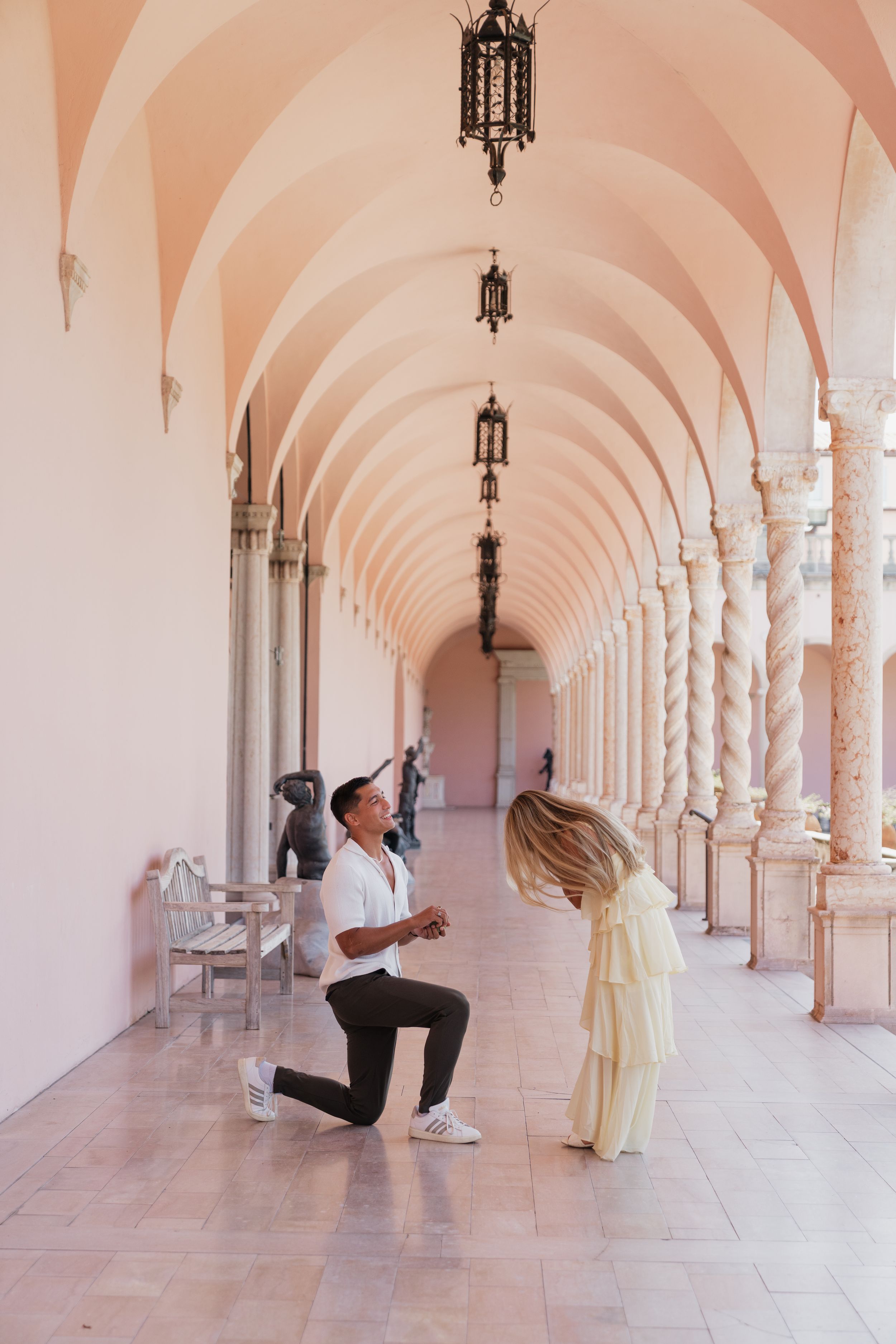 Romantic marriage proposal moment in elegant pink arched hallway with spiral columns.