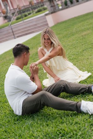 Couple sitting on green grass holding hands, woman in yellow dress smiling at man in white shirt and gray pants.