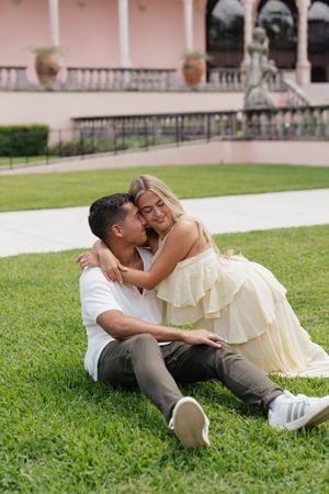Couple sitting on green lawn embracing and smiling near a pink Mediterranean-style building.