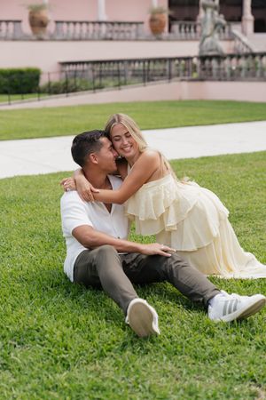 Happy couple sitting together on lawn, woman in yellow dress embracing man in white shirt near elegant building.