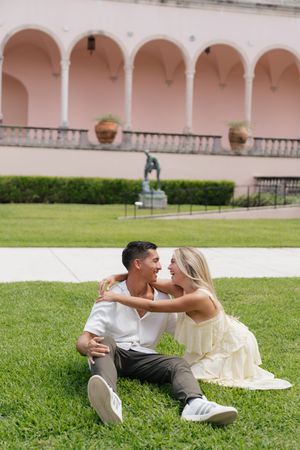 Newly engaged couple sitting on grass embracing in front of pink arched building.