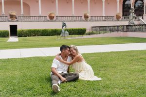 Couple sitting closely together on green lawn in front of pink Mediterranean building.