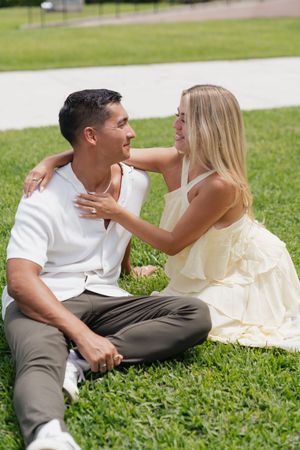 Happy couple sitting on grass, woman in yellow dress embracing man in white shirt during outdoor photo session.