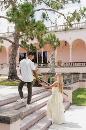 An engaged couple holds hands on elegant stone steps at a pink Mediterranean-style estate with classical statues.